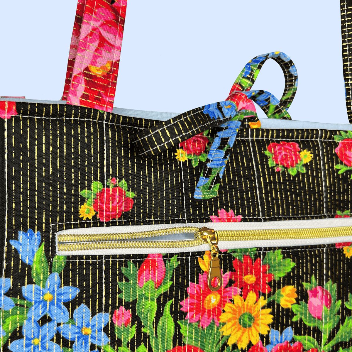 Close-up of a tote bag with a black background, gold vertical stitched lines, and a colorful floral print. The bag features a gold zipper and matching floral shoulder straps.