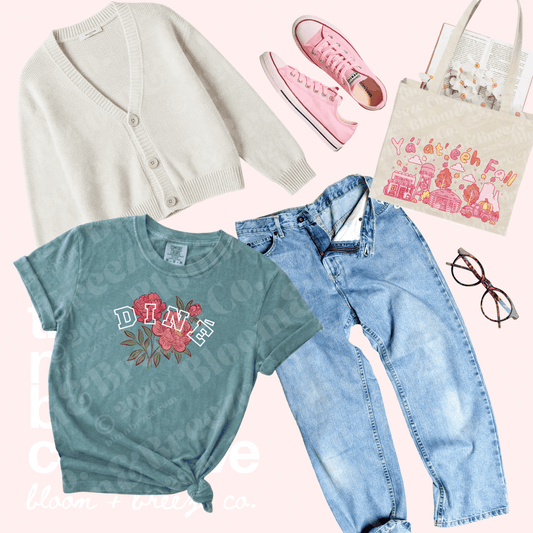 A styled flat-lay outfit arrangement on a pale pink background featuring a teal graphic T-shirt with a floral print and the word 'DINE', a cream cardigan, light-wash blue jeans, pink sneakers, a cream tote bag with a pink illustration, and eyeglasses.