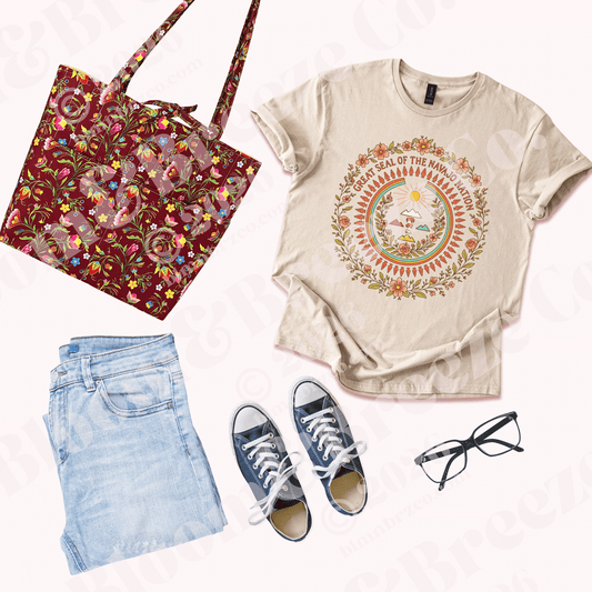 Flat lay outfit with a cream graphic T-shirt featuring a floral Great Seal of the Navajo Nation design, paired with light blue jeans, navy sneakers, black glasses, and a deep berry floral quilted tote bag.