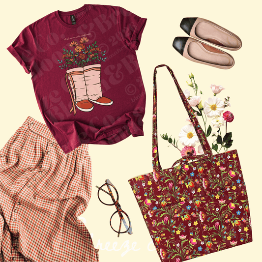 A flat-lay outfit arrangement featuring a burgundy graphic T-shirt with a floral boot print, a matching floral tote bag, a red-and-white gingham shirt, beige flats with black toes, and eyeglasses on a pale cream background.