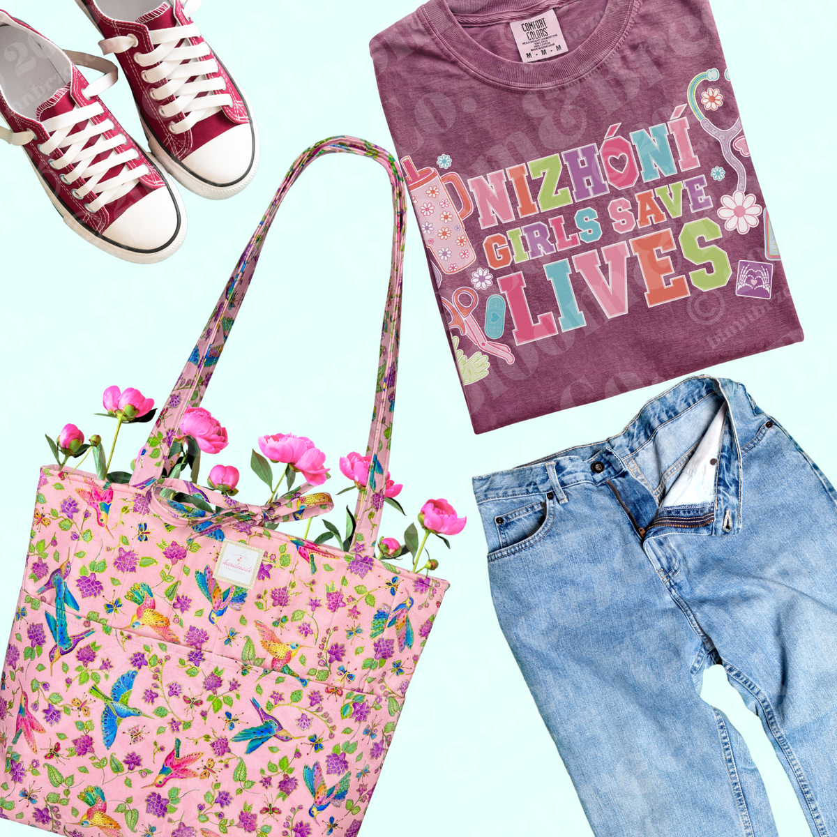 A styled flat-lay featuring a pink floral tote bag with hummingbirds and flowers, a folded purple graphic T-shirt that reads 'NIZHONI GIRLS SAVE LIVES' in colorful letters, a pair of burgundy and white sneakers, and light blue jeans, all arranged on a pale blue background.