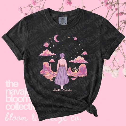 Black t-shirt with a colorful graphic of a woman in a purple dress standing on a planet, on a pink background with floral elements.