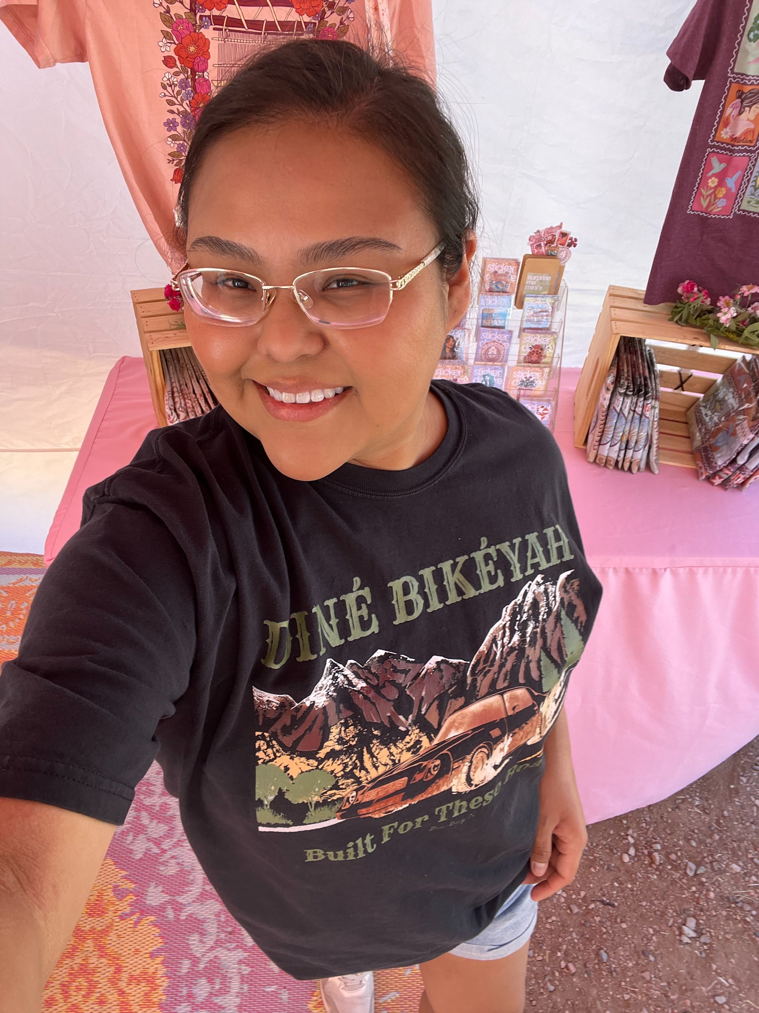 A smiling woman wearing glasses and a black graphic t-shirt with 'dINÉ BIKÉYAH' and a mountain print, taking a selfie in what appears to be a market or booth setting with pink tables and merchandise.