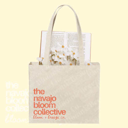 A cream canvas tote bag with orange text reading 'the navajo bloom collective bloom + breeze co.' on a pale yellow background.