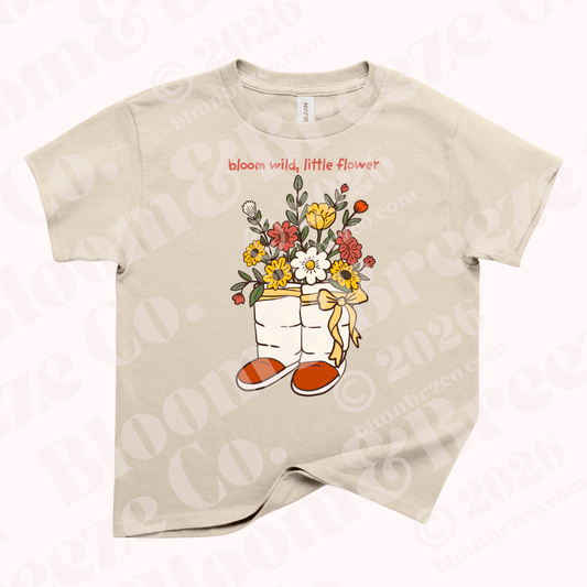 Beige youth t-shirt featuring Navajo wrap moccasins overflowing with yellow, red, and white wildflowers and the phrase “bloom wild, little flower.”