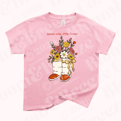 Pink youth t-shirt with illustration of Navajo wrap moccasins filled with colorful wildflowers and the words “bloom wild, little flower” above the design.