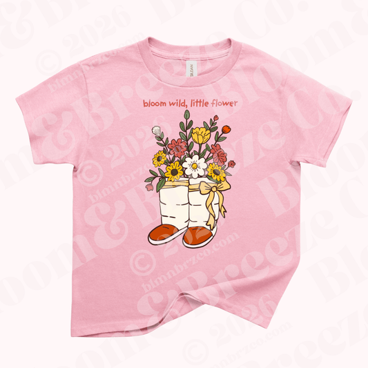 Pink youth t-shirt with illustration of Navajo wrap moccasins filled with colorful wildflowers and the words “bloom wild, little flower” above the design.