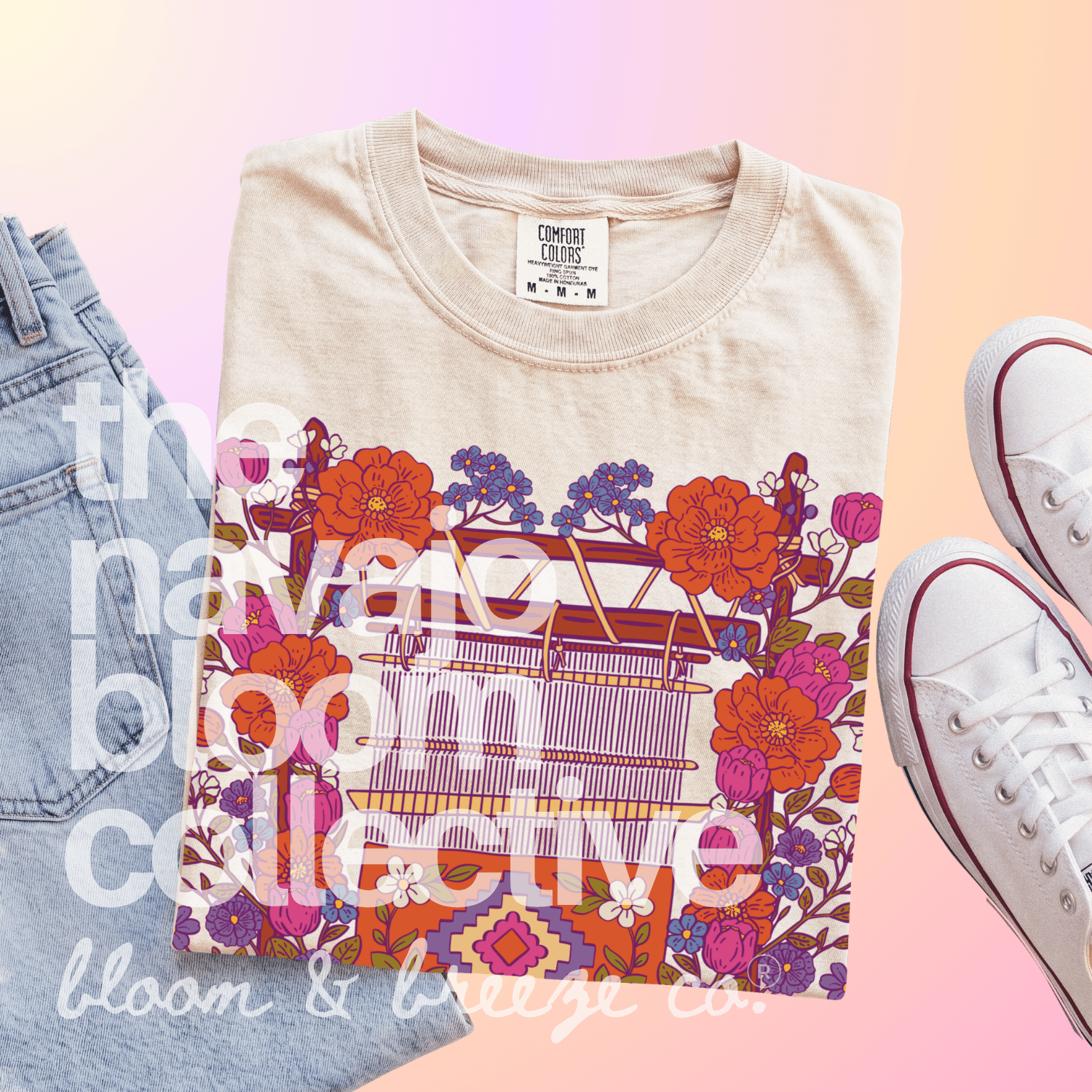 A folded beige graphic T-shirt is styled in a flat lay with light blue jeans on the left and white low-top sneakers on the right, set against a soft pink-to-peach gradient background. The shirt features a colorful floral and geometric pattern with a sewing machine illustration.