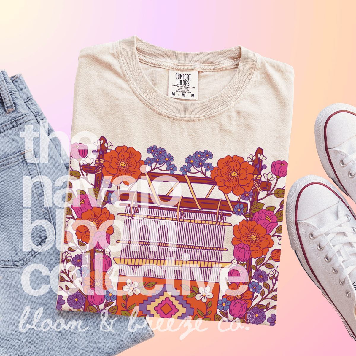 A folded beige graphic T-shirt is styled in a flat lay with light blue jeans on the left and white low-top sneakers on the right, set against a soft pink-to-peach gradient background. The shirt features a colorful floral and geometric pattern with a sewing machine illustration.