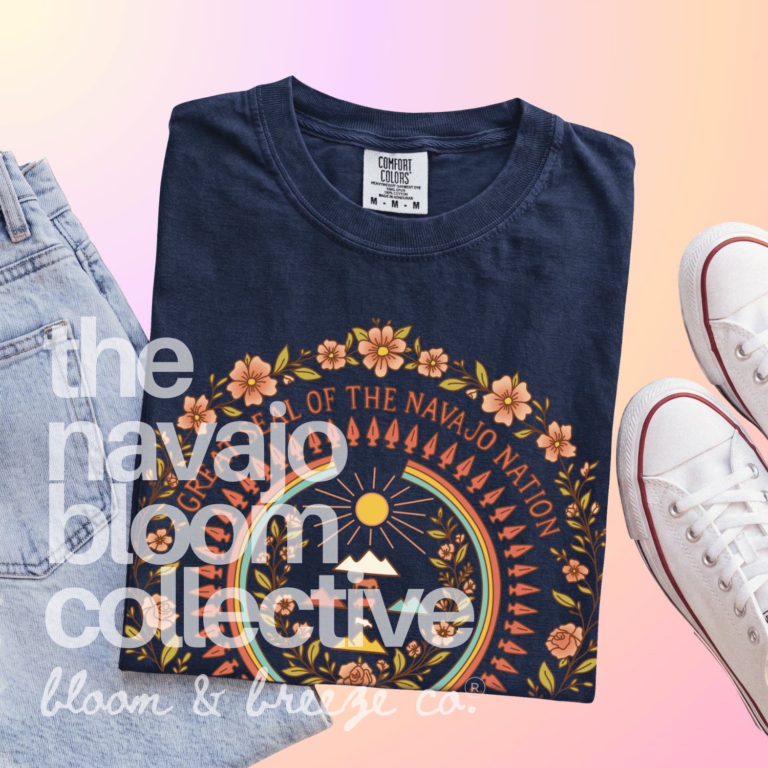 A folded navy blue graphic T-shirt is displayed on a pastel pink-to-yellow gradient background, styled with light-wash jeans on the left and white sneakers on the right. The shirt features a large circular Native American-inspired design with orange flowers, a rainbow, and a sun, with text reading 'the navaio bloom collective' and 'bloom & breeze co.