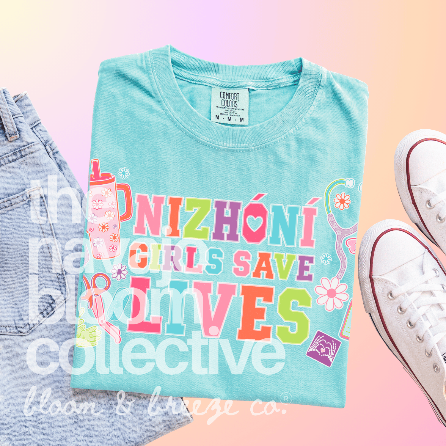 A folded light blue graphic T-shirt is displayed on a pastel pink-to-yellow gradient background, styled with part of a denim jacket on the left and white sneakers on the right. The shirt features the text 'NIZHÓNÍ GIRLS SAVE LIVES' in large multicolored letters with small decorative illustrations including flowers, a pink water bottle, and a ribbon.