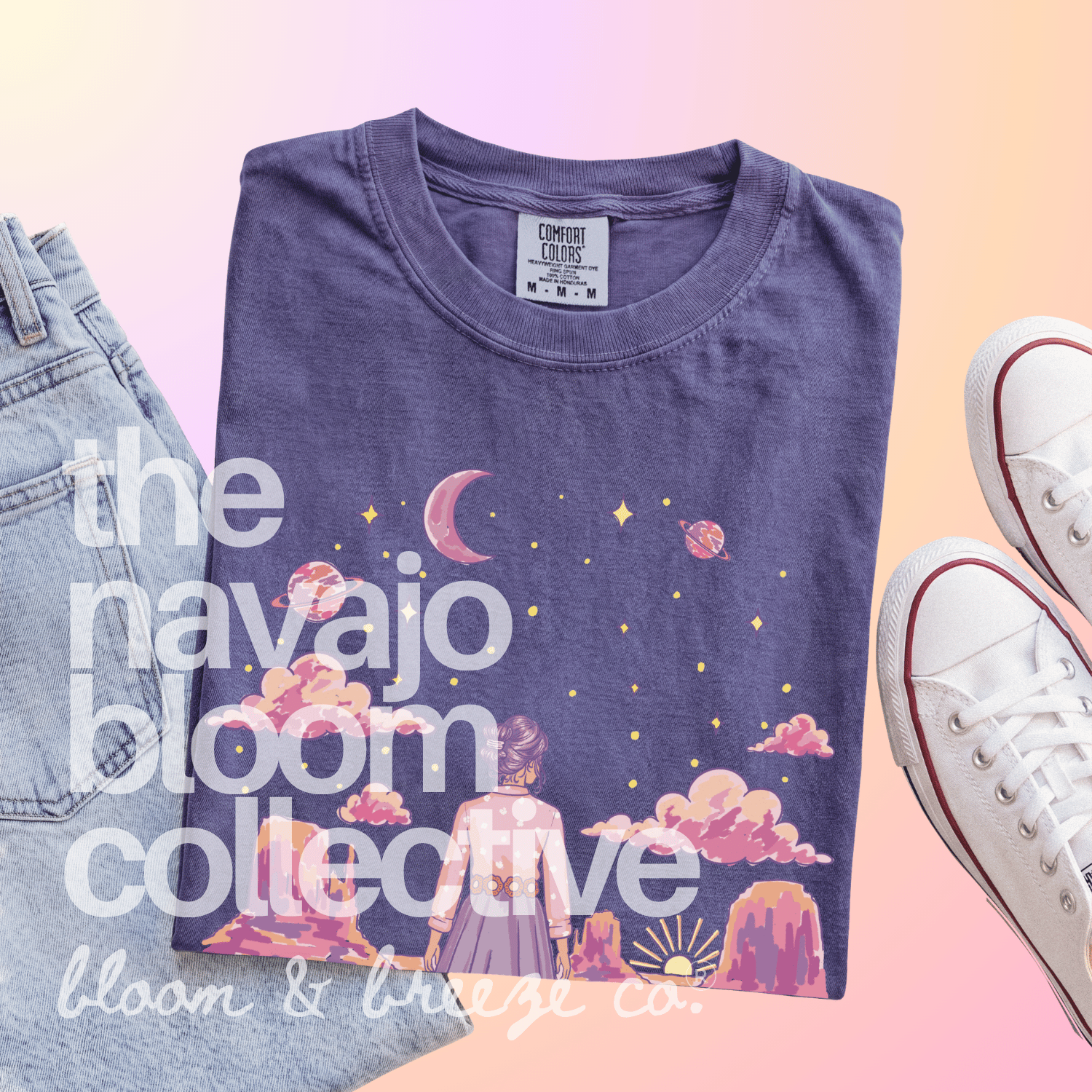 A folded purple graphic T-shirt is styled in a flat lay with light-wash jeans on the left and white sneakers on the right, set against a soft pink-to-yellow gradient background. The shirt features a whimsical celestial print with a girl, mountains, and stars.
