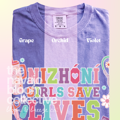 Nizhóní Girls Save Lives colorful lettering and medical-themed artwork on a Comfort Colors tee, comparing Grape, Orchid, and Violet.