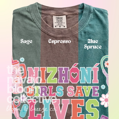 Nizhóní Girls Save Lives colorful lettering and medical-themed artwork on a Comfort Colors tee, comparing Sage, Espresso, and Blue Spruce.