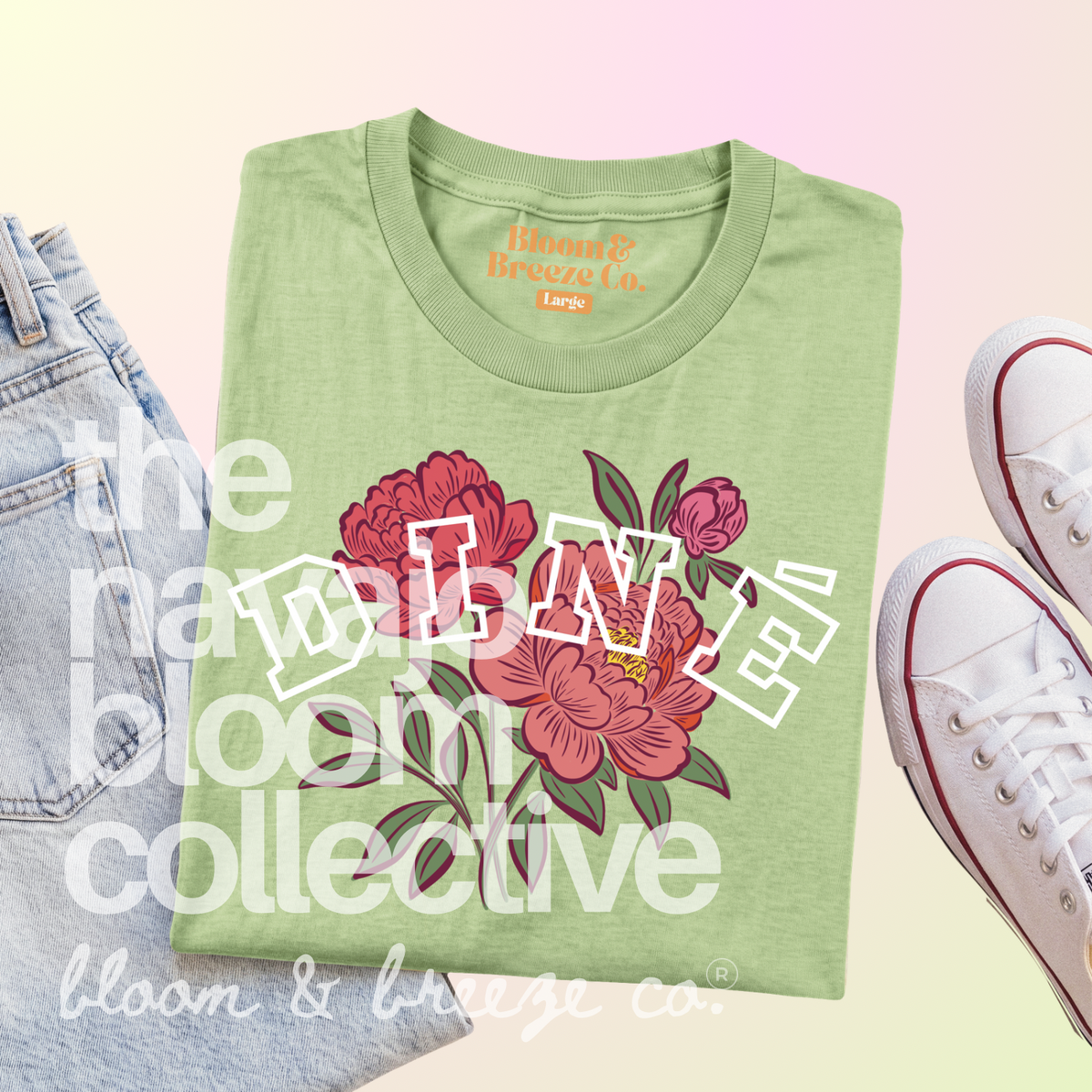 A folded sage green graphic T-shirt from Bloom & Breeze Co. is styled in a flat lay with light-wash jeans on the left and white sneakers on the right.