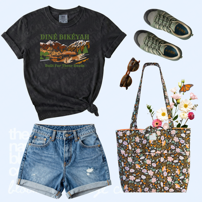 Black Diné Bikéyah graphic tee styled with denim shorts, green sneakers, sunglasses, and a dark floral quilted tote bag.