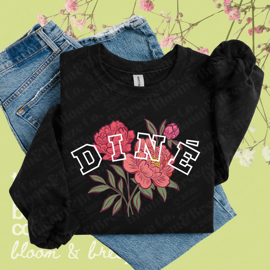 Black Diné floral sweatshirt layered over denim jeans on a soft green floral background.