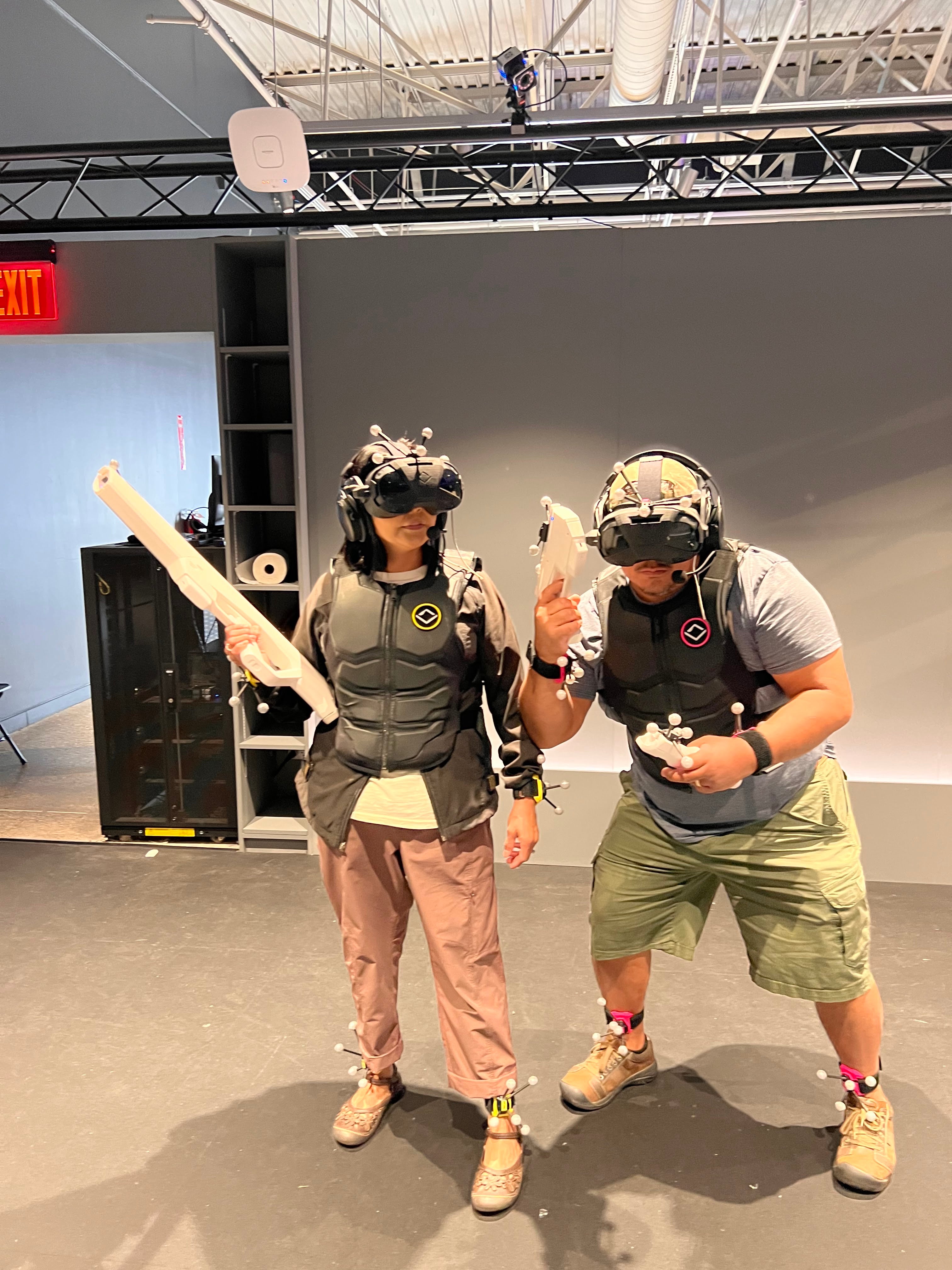 Two people wearing VR headsets and protective vests engage in a virtual reality combat simulation inside an indoor training facility, holding foam weapons.
