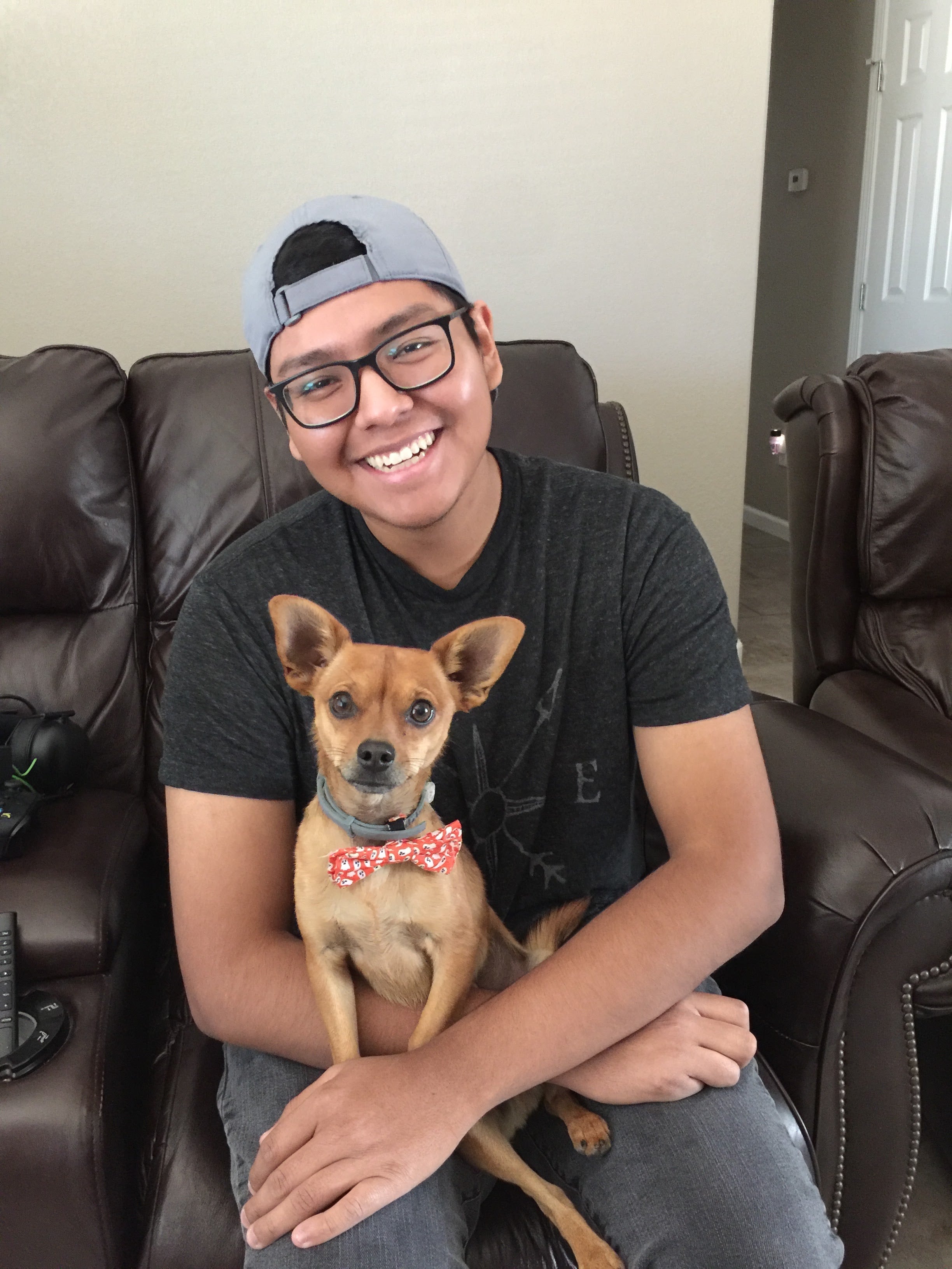 A smiling man wearing a gray cap and glasses sits on a brown leather couch holding a small tan dog with a red bow tie.