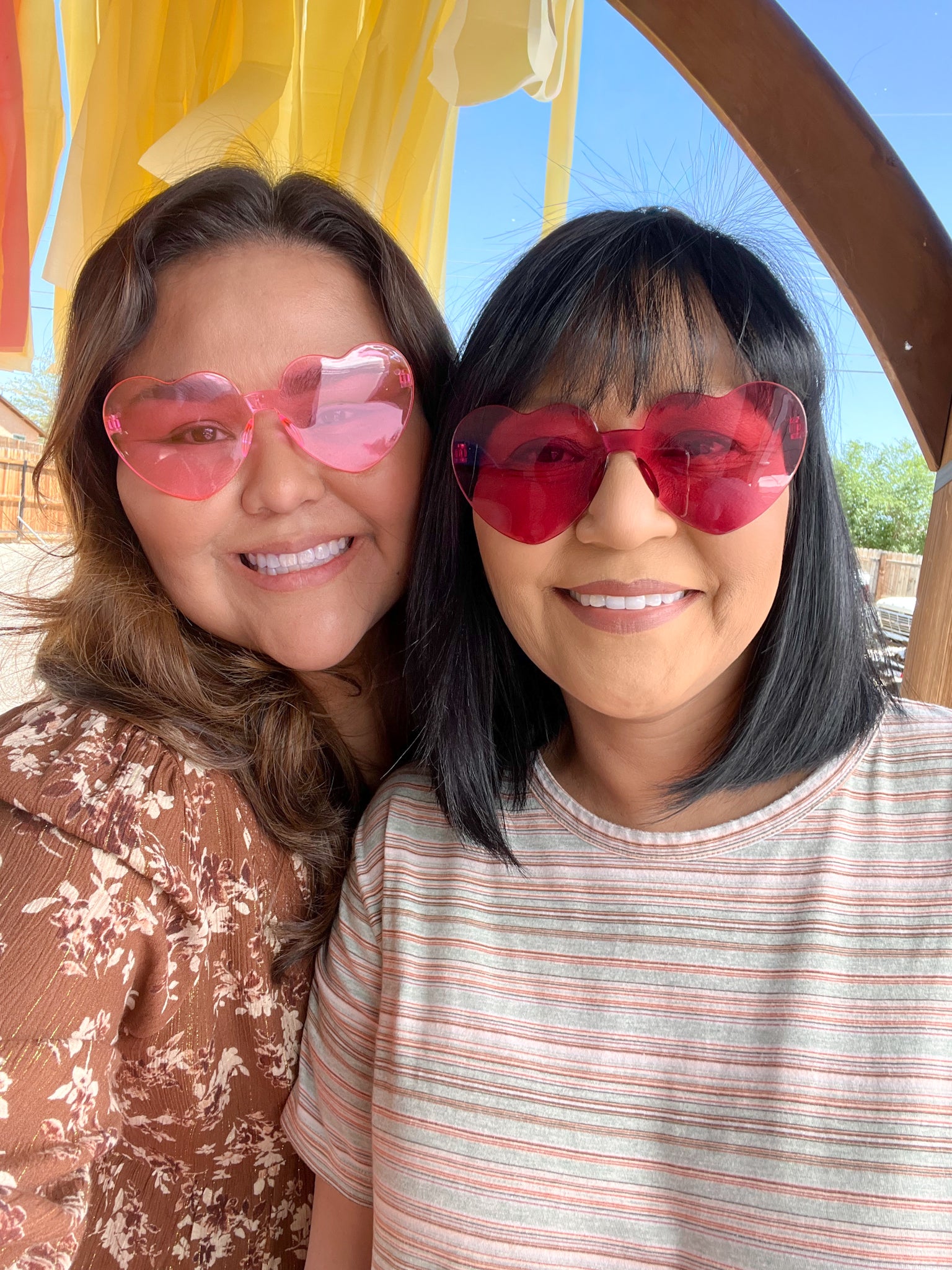 Two women smiling and taking a selfie outdoors under a wooden arch with yellow fabric. Both are wearing heart-shaped sunglasses with pink and red lenses.