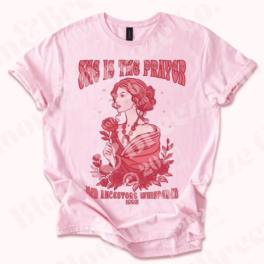 Pink graphic t-shirt reading “She Is the Prayer – Her Ancestors Whispered 1993” featuring a woman holding a flower surrounded by red florals.