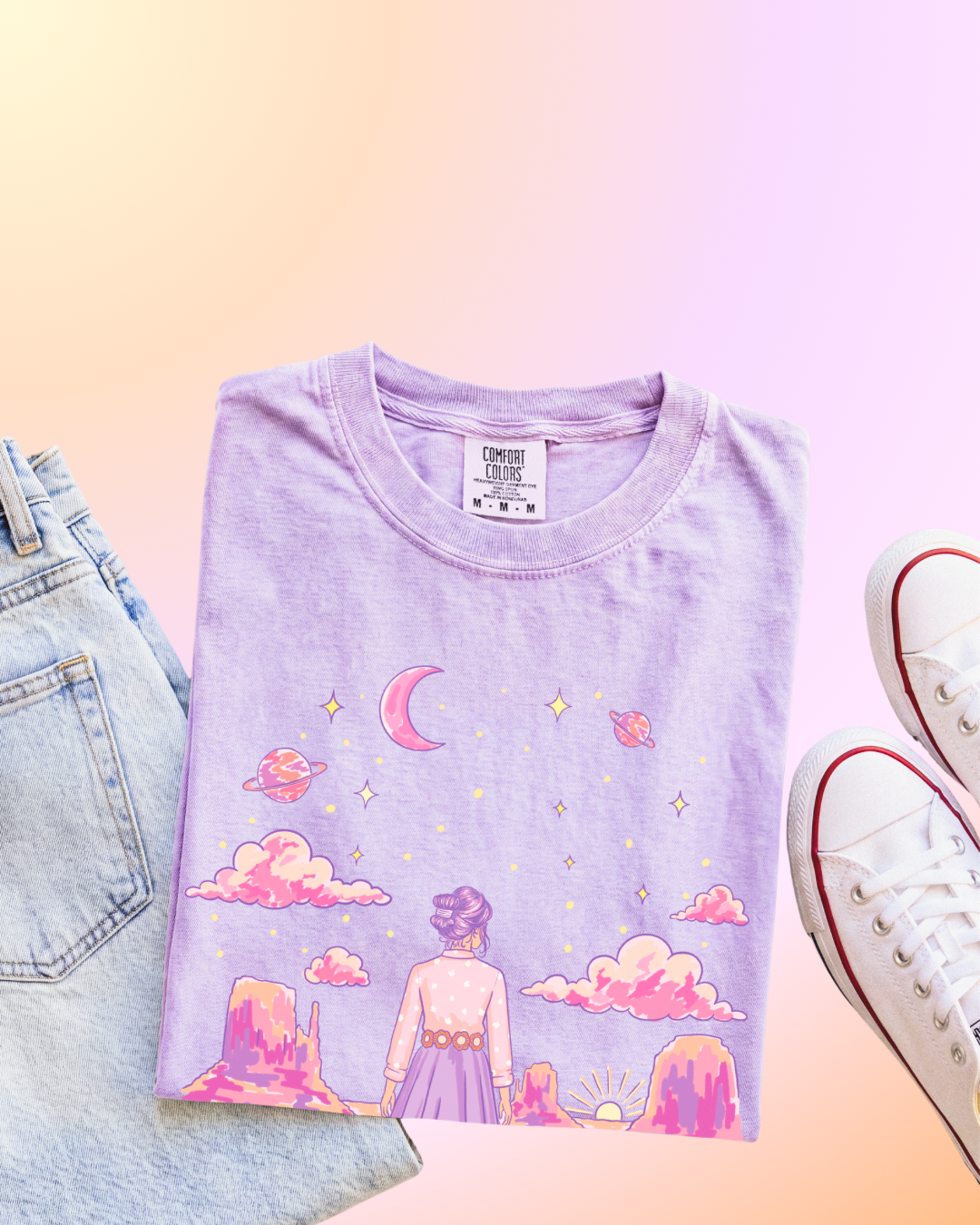 A folded lavender t-shirt with a colorful pastel graphic of a girl looking at a starry sky, styled in a flat lay with light-wash jeans and white sneakers on a pastel gradient background.