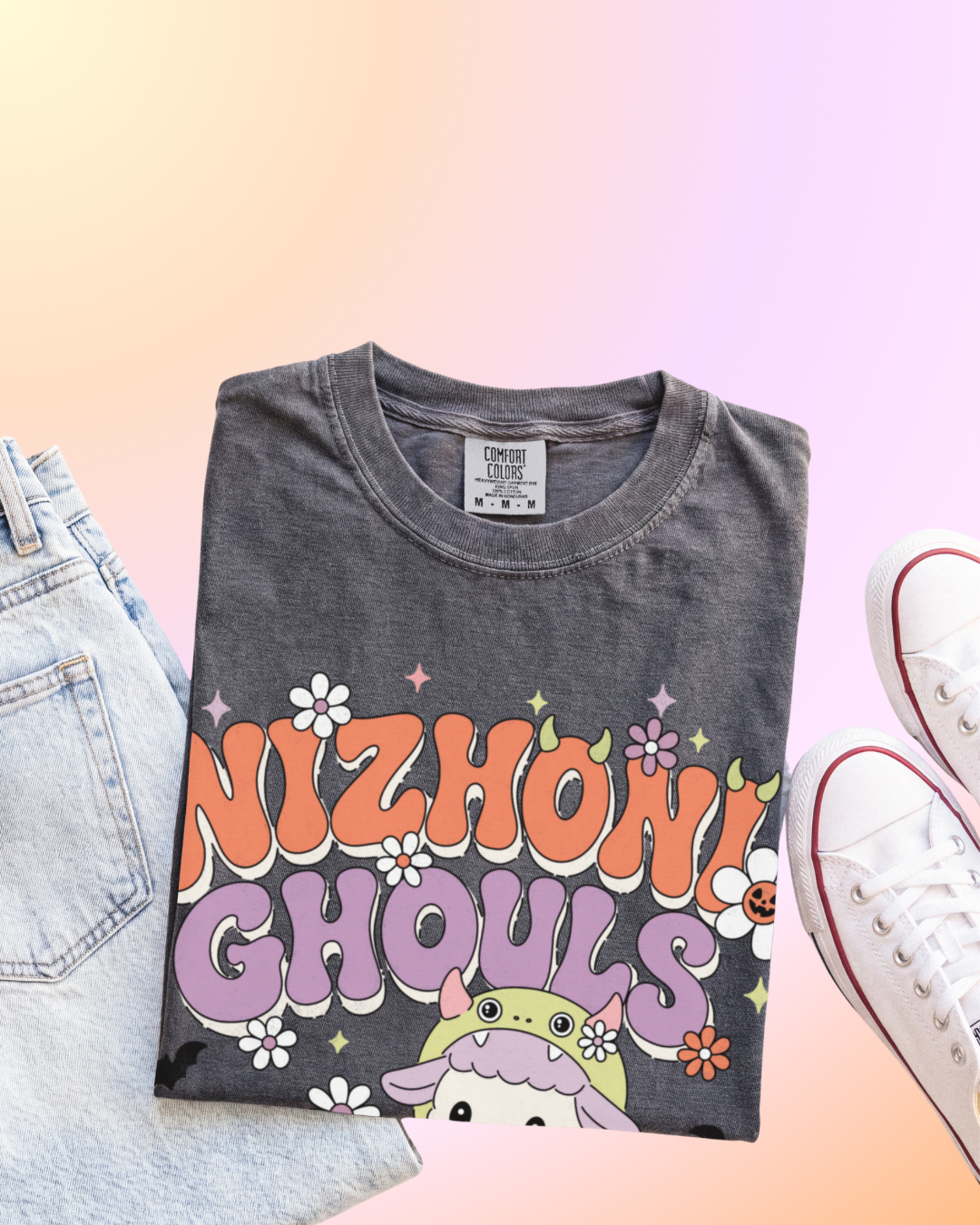 A folded dark gray graphic t-shirt with 'NIZHONI GHOULS' in retro orange, purple, and white lettering, styled in a flat-lay with light blue jeans and white sneakers on a pastel gradient background.