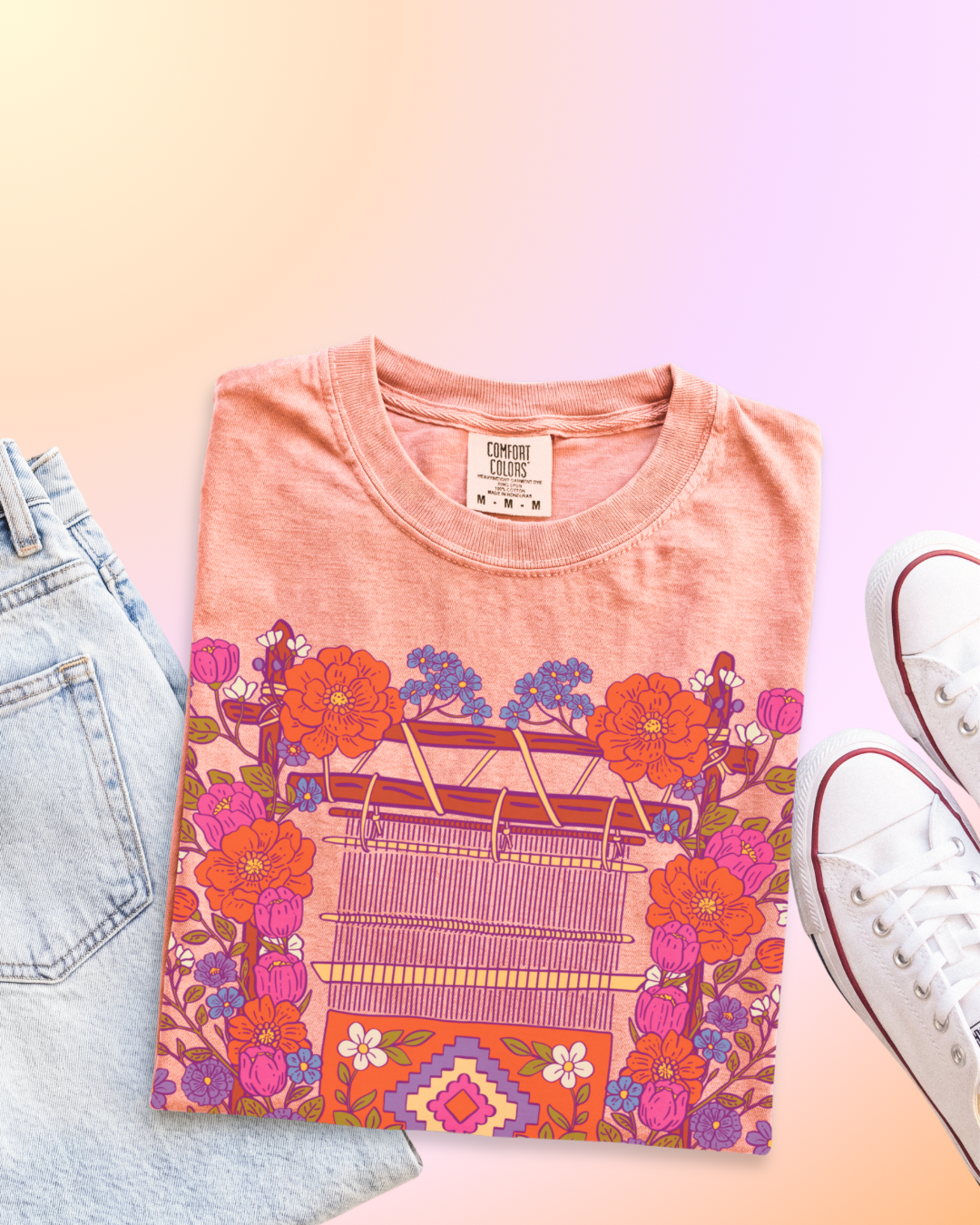 A folded peach-pink t-shirt with a colorful retro-style graphic of a loom, flowers, and cartoon characters, styled in a flat lay with light-wash jeans and white sneakers on a pastel gradient background.