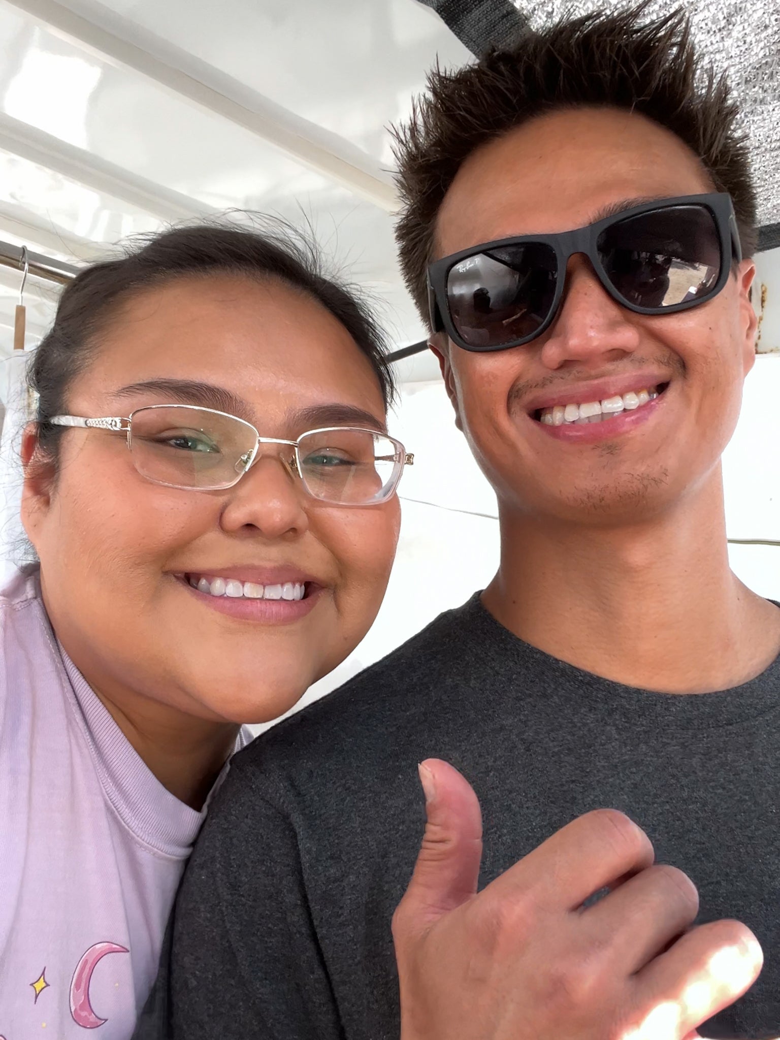 A close-up selfie of a smiling couple. The woman on the left wears clear-framed glasses and a light lavender top with a moon graphic.