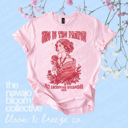 Pink “She Is the Prayer” graphic tee with red illustrated woman and floral design on a soft blue floral background.