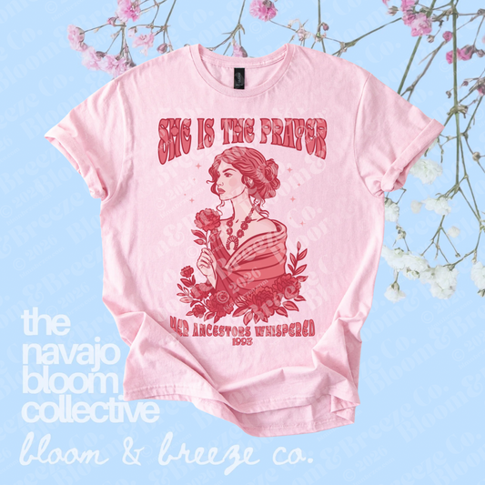 Pink “She Is the Prayer” graphic tee with red illustrated woman and floral design on a soft blue floral background.