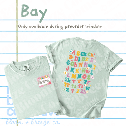 A light whitish green graphic t-shirt shown folded and flat, with text overlays reading 'Bay' and 'Only available during preorder window' on a lined paper background. The shirt features colorful alphabet letters with small floral accents on the back.