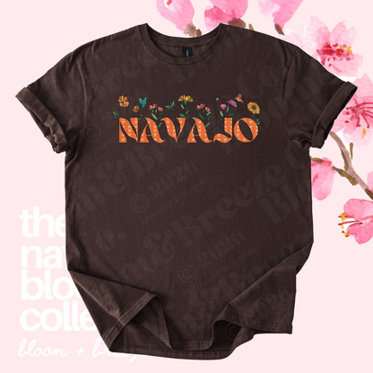 A dark brown short-sleeve t-shirt with a graphic on the chest featuring the word 'NAVajo' in orange letters filled with a floral pattern, surrounded by small multicolored flowers. The shirt is displayed flat against a light pink background decorated with cherry blossom graphics and faint text watermarks.