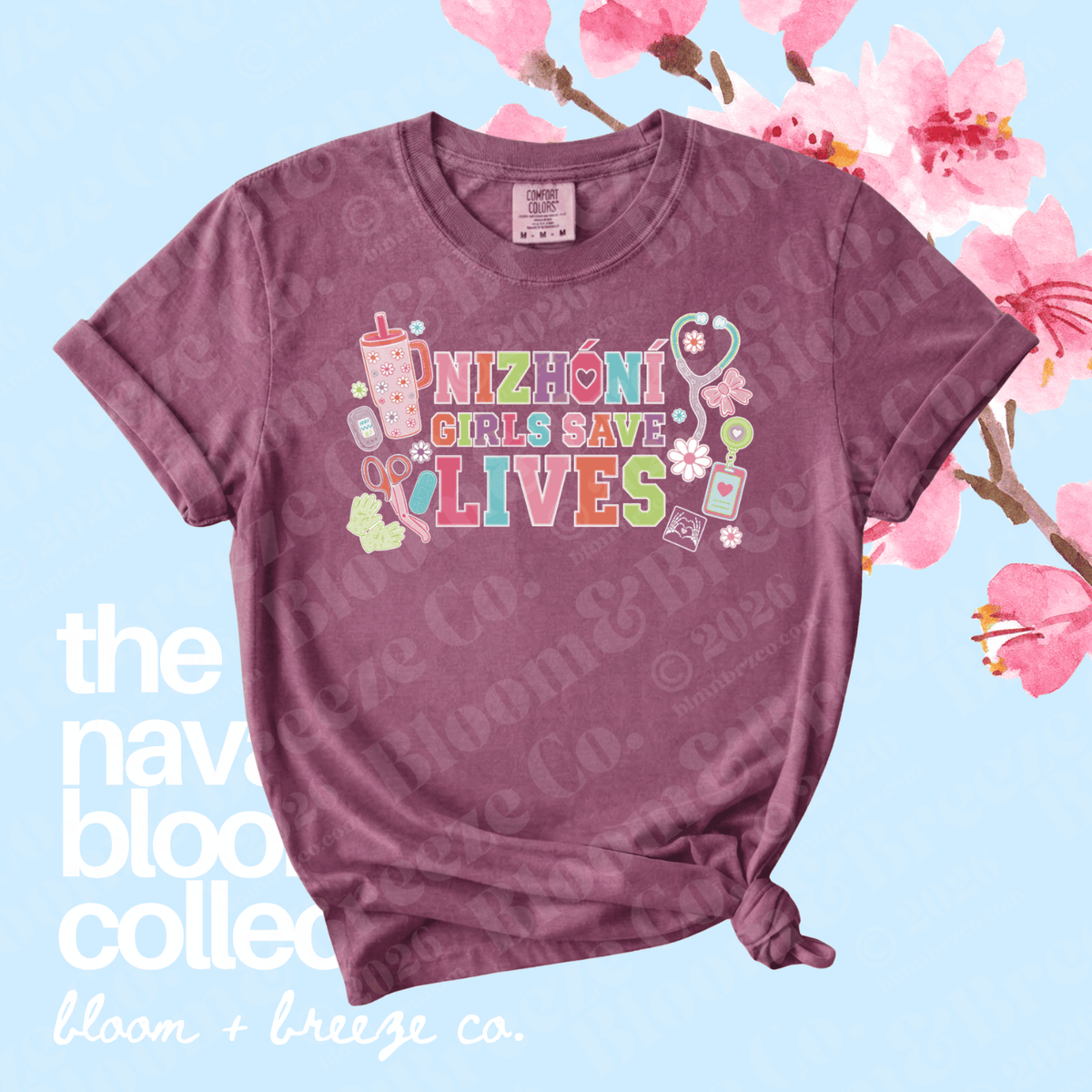 A mauve short-sleeve graphic T-shirt with rolled cuffs and a tied hem, featuring the phrase 'NIZHONI GIRLS SAVE LIVES' in colorful lettering with medical-themed illustrations. The shirt is displayed on a light blue background with pink cherry blossom branches and text overlays reading 'the nav blood collection' and 'bloom + breeze co.