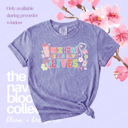 A lavender heathered graphic t-shirt with rolled sleeves and a knotted hem, featuring the text 'NIZHONI GIRLS SAVE LIVES' in colorful letters with medical-themed illustrations. The shirt is displayed on a light lavender background decorated with pink cherry blossoms and text overlays.