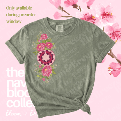 A short-sleeve crewneck t-shirt in a muted sage green, featuring a vertical graphic print of pink flowers and a central pink-and-white geometric heart design. The shirt is styled with a knot at the bottom hem against a light pink background decorated with cherry blossoms and text overlays.