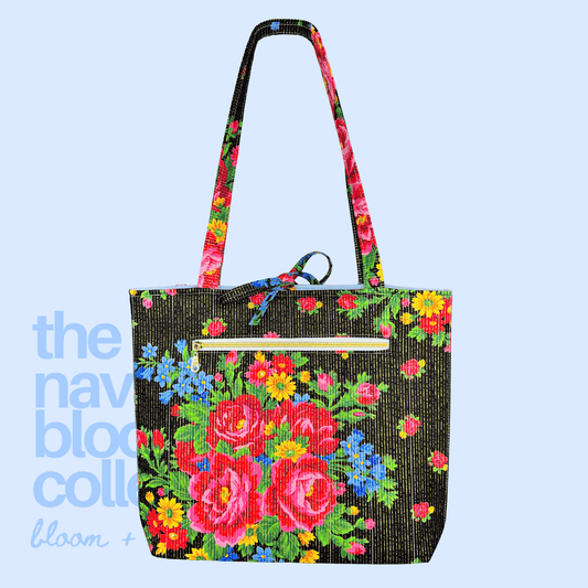 Back view of a black floral tote with vivid red, blue, and yellow flowers, long shoulder straps, and a gold-zippered exterior pocket.