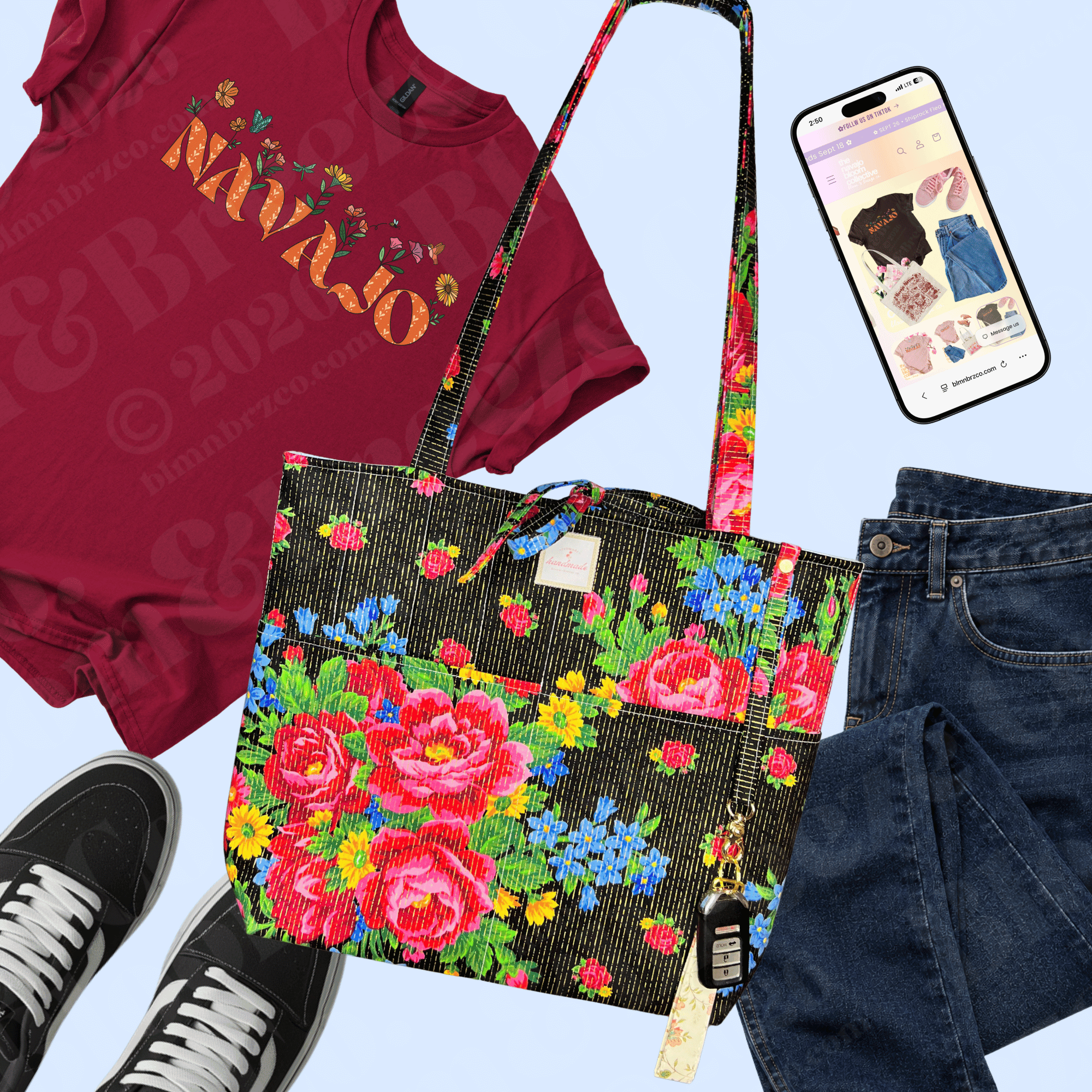 Flat lay of a maroon “Navajo” floral T-shirt styled with dark jeans, black sneakers, a black floral tote, car keys, and a smartphone. ⠋