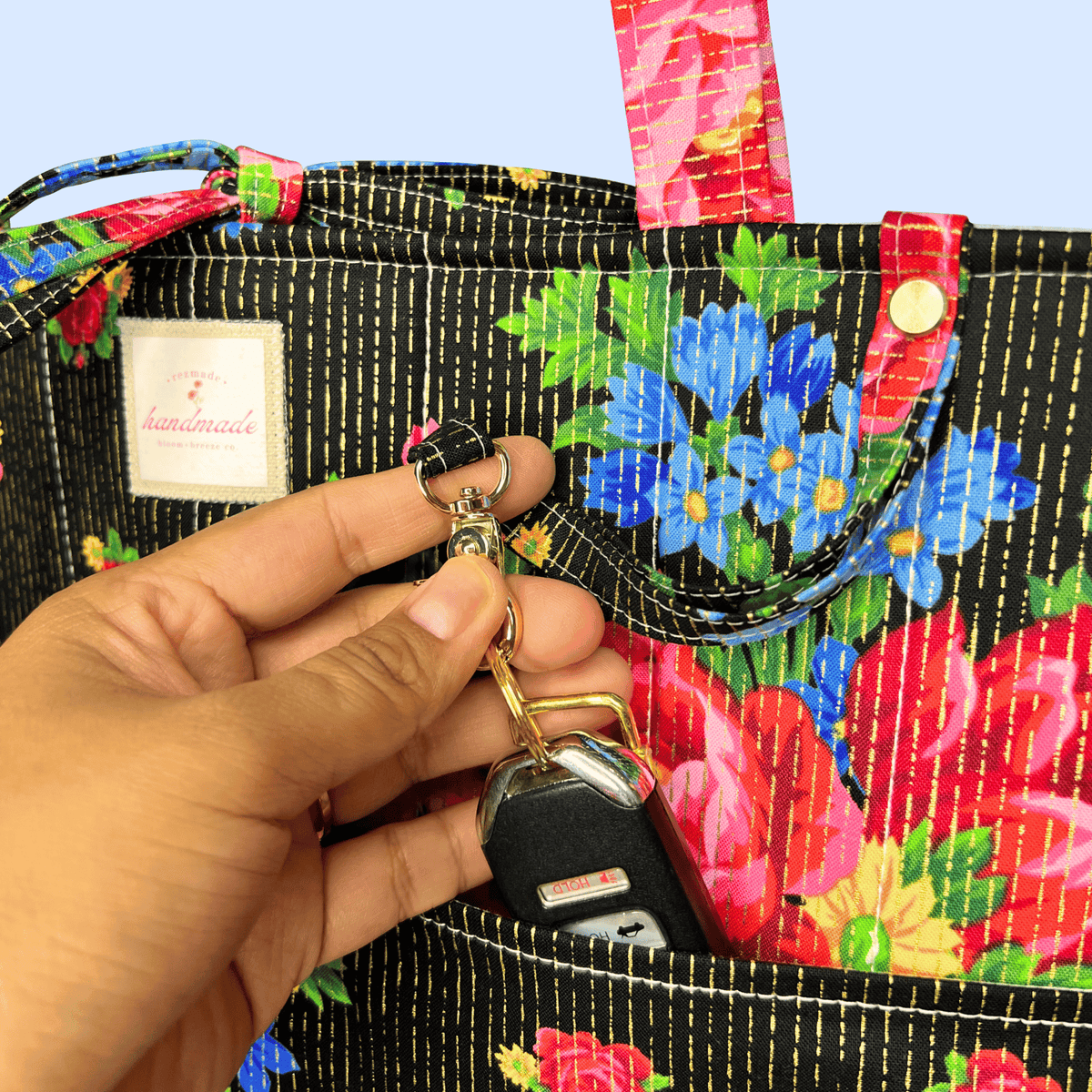 Close-up of a black tote with colorful red and blue flowers and gold stitching as a hand clips car keys to an exterior ring.