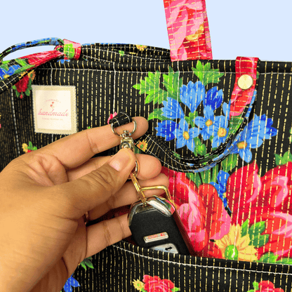 Close-up of a black tote with colorful red and blue flowers and gold stitching as a hand clips car keys to an exterior ring.