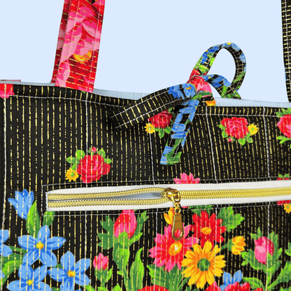 Close-up of a tote bag with a black background, gold vertical stitched lines, and a colorful floral print. The bag features a gold zipper and matching floral shoulder straps.