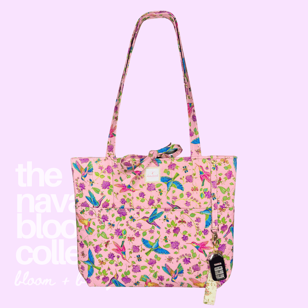 A pink tote bag with a vibrant multicolor floral and bird print, featuring a matching shoulder strap, a front pocket, and a hanging paper tag, set against a solid light purple background with white text overlay.