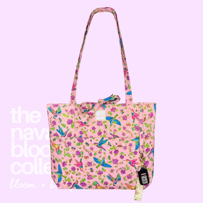A pink tote bag with a vibrant multicolor floral and bird print, featuring a matching shoulder strap, a front pocket, and a hanging paper tag, set against a solid light purple background with white text overlay.