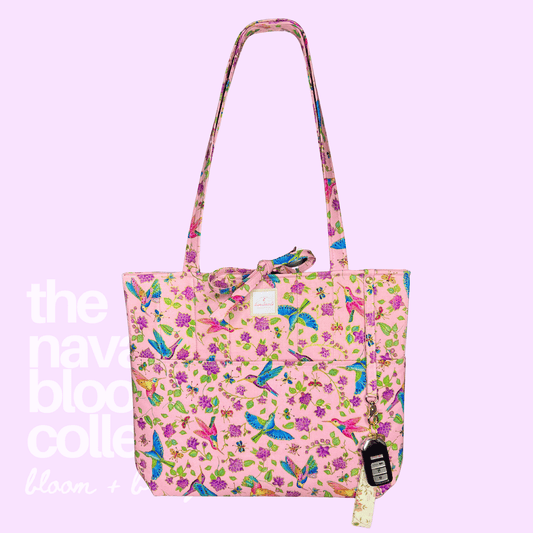 A pink tote bag with a vibrant multicolor floral and bird print, featuring a matching shoulder strap, a front pocket, and a hanging paper tag, set against a solid light purple background with white text overlay.