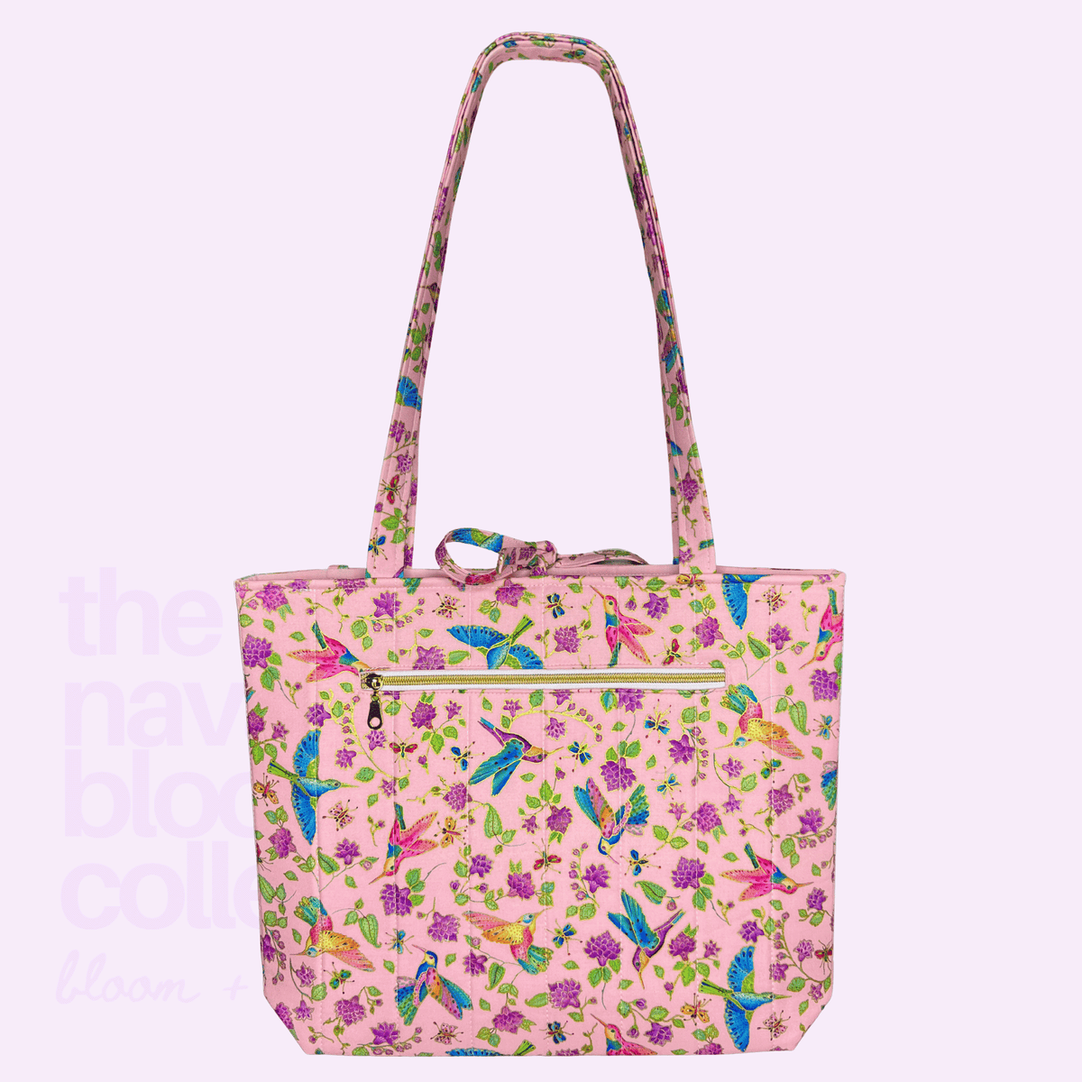 Back view of a pink hummingbird, butterfly, and floral-print tote with long shoulder straps and a gold-zippered exterior pocket.