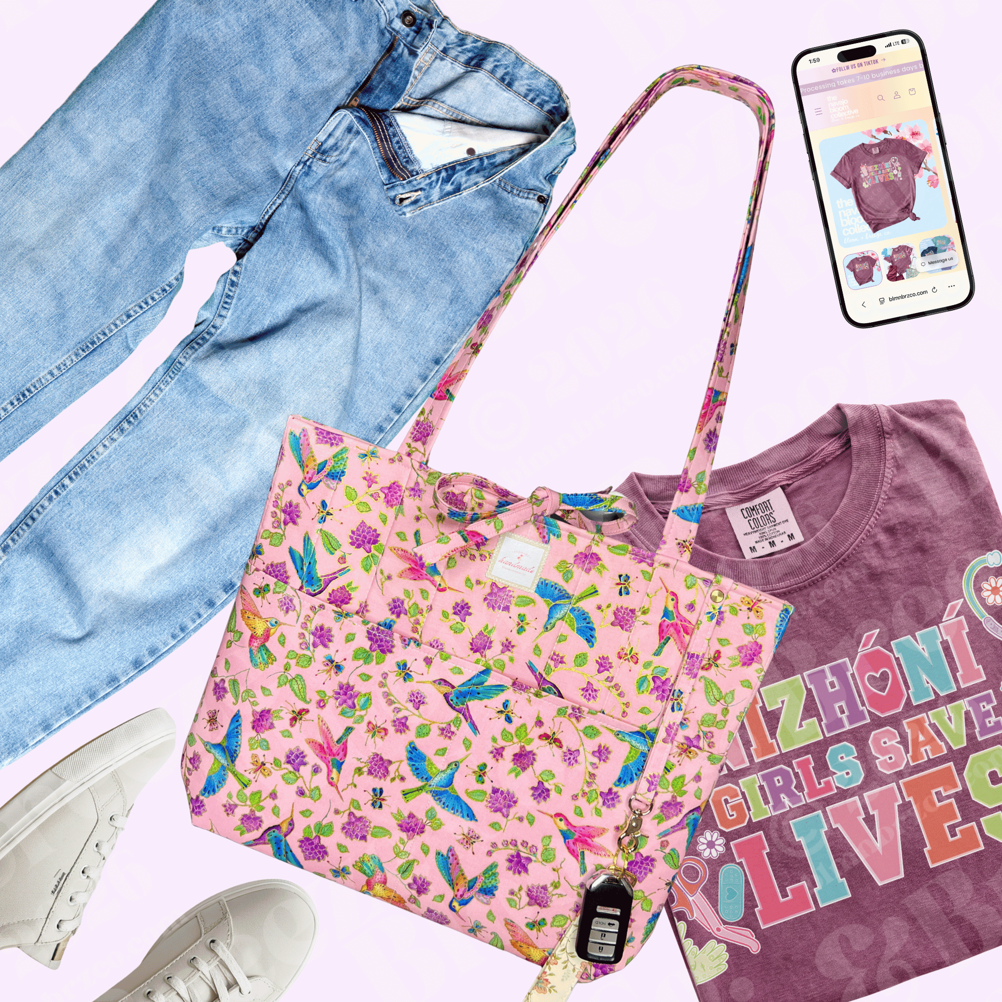 Flat lay of a mauve “Nizhóní Girls Save Lives” T-shirt styled with light-wash jeans, white sneakers, a pink hummingbird-print tote, car keys, and a smartphone.