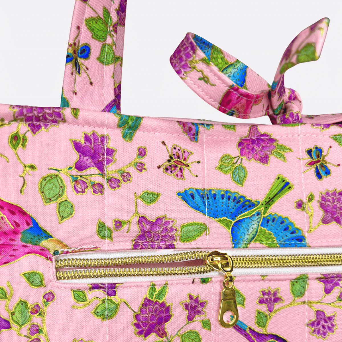Close-up of a pink hummingbird, butterfly, and floral-print tote featuring a gold-zippered exterior pocket and tied handles.