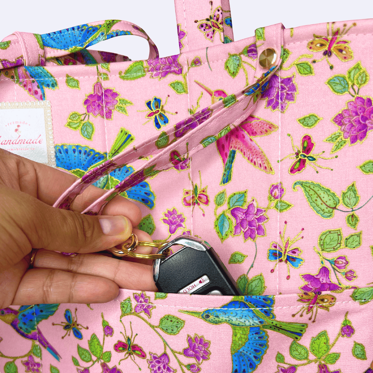 Close-up of a pink hummingbird, butterfly, and floral-print tote as a hand places car keys into the front pocket.