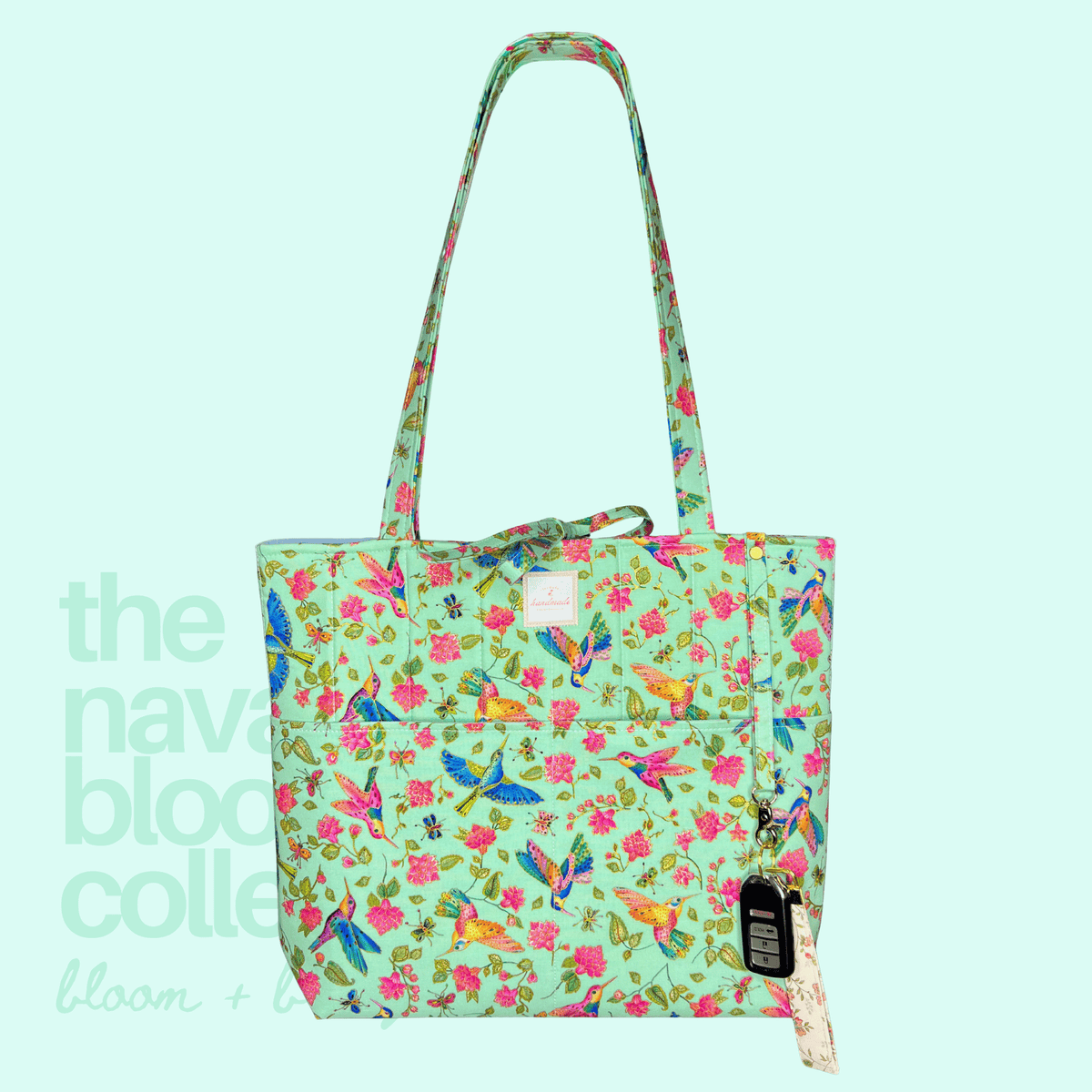A light green tote bag with a vibrant multicolor floral and bird print, featuring long shoulder straps, a front pocket, and a hanging fabric tag, set against a pale mint green background with a brand watermark.