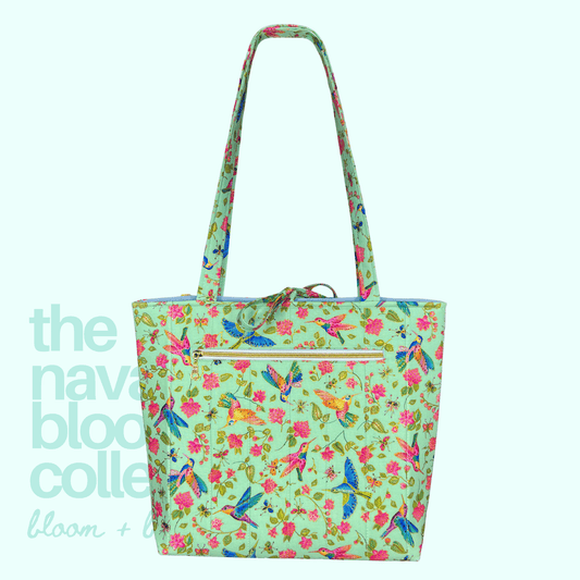 A tote bag with a light mint green background and a colorful print of hummingbirds, butterflies, and pink flowers. It features dual shoulder straps, a front zippered pocket, and a drawstring detail at the top.